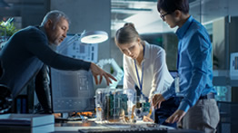 A supervisor reviews a project with two staff members centered around a computing device placed on a table